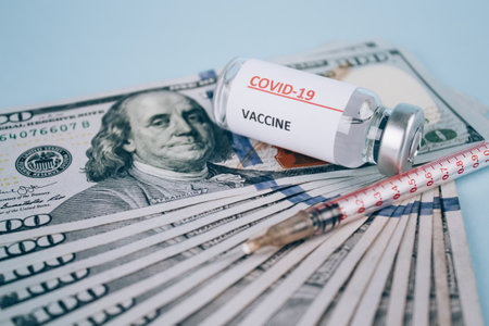 Dollars bills and vial dose of COVID-19 vaccine with syringe against blue background with copy space - global vaccination and money abuse concept. Covid vaccine cost, selective focusの写真素材