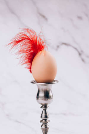 Trendy still life Easter composition with hen egg in vintage candlestick with bright red feather against light marble background. Easter egg as a symbol of universe, resurrection and circle of lifeの写真素材