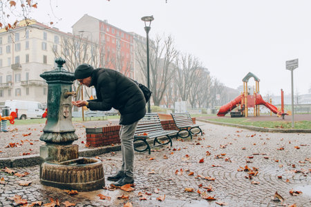 Milan, Italy - January 1, 2018 : Tourist using Vedovelle - typical dark green cast-iron city drinking fountain in Milan, public water supply and drinking water safety concept. Travel tipsのeditorial素材