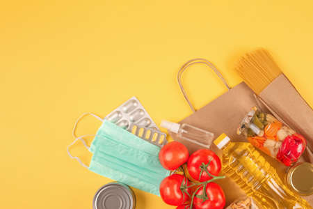Food and medicines donations on yellow background with copy space - pasta, vegatables, canned food, cooking oil, hand sanitizer and face masks. Food bank concept. Selective focus, flat layの写真素材