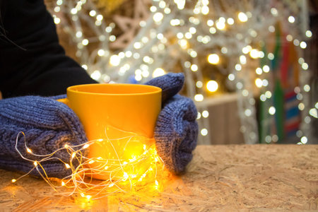 Woman's hands holding yellow mug with hot tea or coffee with illumination lights around. Christmas...