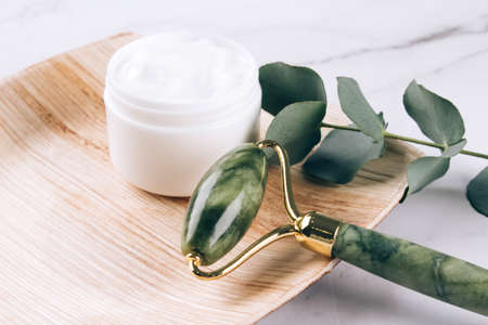Jade facial gua sha roller with moisturizing face cream on white marble table background with copy space. Facial massage kit for lifting massage therapy made from natural stones, mock upの写真素材