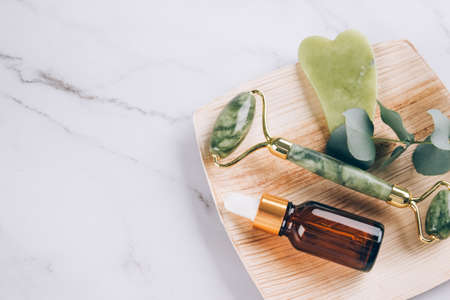 Jade gua sha facial roller with beauty serum oil on white marble table background with copy space. Facial massage kit - for lifting massage therapy made from natural stones. Selective focusの写真素材