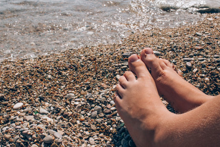 Unrecognisable child sitting on the beach near water, children's legs against sea shore as summer...
