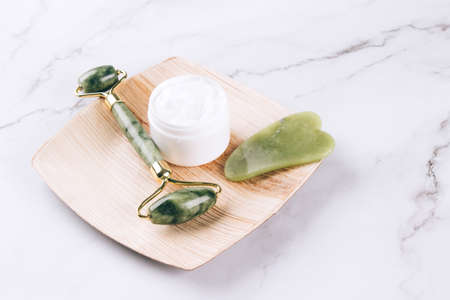 Jade facial gua sha roller with moisturizing face cream on white marble table background with copy space. Facial massage kit for lifting massage therapy. Mock up. Selective focusの写真素材