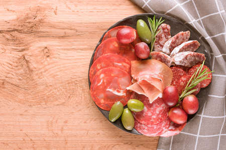 Top view on charcuterie plate with different types of sausages - salami, bresaola, proscuitto served with olives and grape. Black plate with traditional italian antipasti on wooden table background with copy space. Selective focusの写真素材