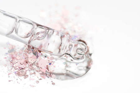 Piipette with transparent liquid on light glass background with splash of shiny pink glitter around and copy space. Texture of face serum or cosmetic oil. Selective focus, macro photoの写真素材