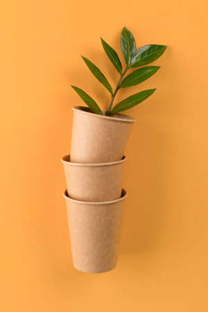 Vertical mockup image of kraft paper coffee cups with green leaves inside - biodegradable, compostable paper utensils for hot drinks. Paper cup on orange background with copy space. Selective focusの写真素材