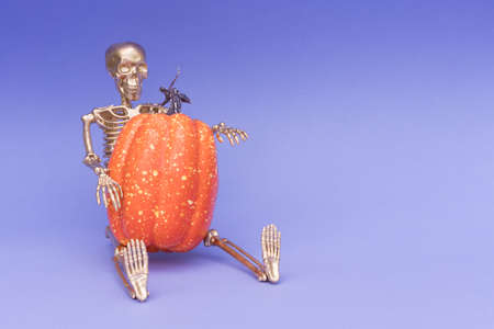 Funny halloween composition with golden skeleton sitting with big orange pumpkin over purple background with copy space for text. Party invitation templateの写真素材