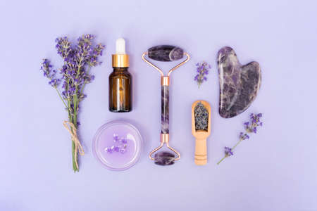 Amethyst gua sha roller and scraper for facial massage with bottle of face serum or lavender cosmetic oil and moisturizer over purple background. Facial treatments kit and skincare conceptの写真素材
