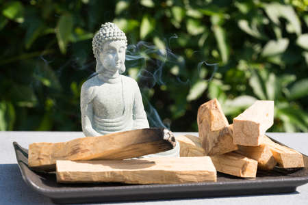 Incense burning. Burning palo santo sticks with Buddha statue against green leaves as background under the sunlight. Zen concept, meditation and spiritual practices. Selective focus, focus on statueの写真素材