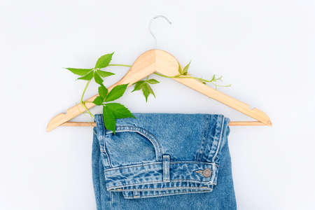 Upcycling clothes or creative reuse concept. Jeans on wooden hanger with green leaves over light gray background. Eco-friendly shopping and slow fashionの写真素材