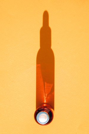 View above glass dropper bottle with face serum over orange background with bottle shape shadow....