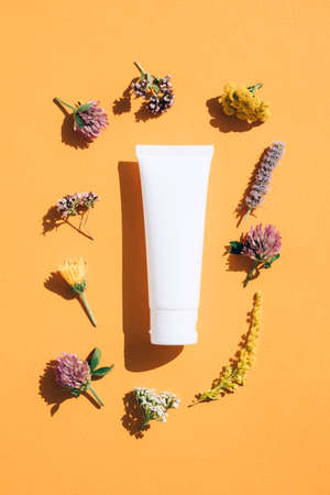 Herbal beauty products - tube with hand cream, face cream or body lotion with natural ingredients with flowers around over orange background. Flat lay style, mockup. Vertical imageの写真素材