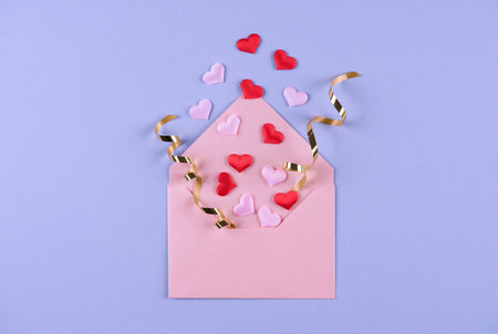 Pink envelope with red hearts confetti and golden ribbons around over purple background. Valentine's day festive background. Flat lay style. Greeting card templateの写真素材