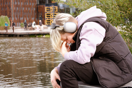 Depressed caucasian teenager boy sitting on the lake shore and listening to music on wireless headphones and crying. Teenage issues and problems conceptの写真素材