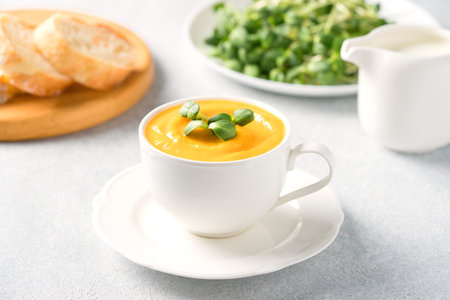 Cup of pumpkin cream soup garnished with fresh sunflower microgreen. Creamy butternut squash autumn soup in elegant white tea cup.  Front view, selective focusの写真素材