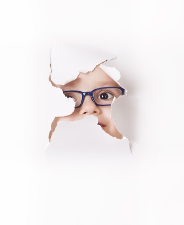 Curious kid in spectacles looks through a hole in white paperの写真素材