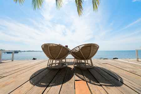 Two wooden deck chairs with nobody in them on a wooden deck overlooking tropical blue cloudy sky and sea.の写真素材