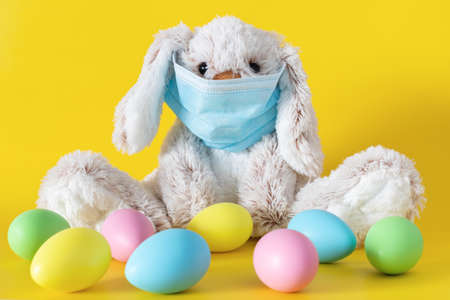 Easter stuffed toy bunny in protective face mask and colored eggs on yellow background. Ester holiday during  pandemic concept.の写真素材
