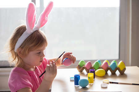 Little girl in Easter bunny ears painting colored eggs. Easter family holiday celebration at home...