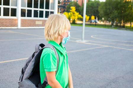 Kid in mask during virus outbreak. Little schoolboy wears grey backpack, breathes through mask, going to school. Back to school concept after reopening. Student at the school yardの写真素材