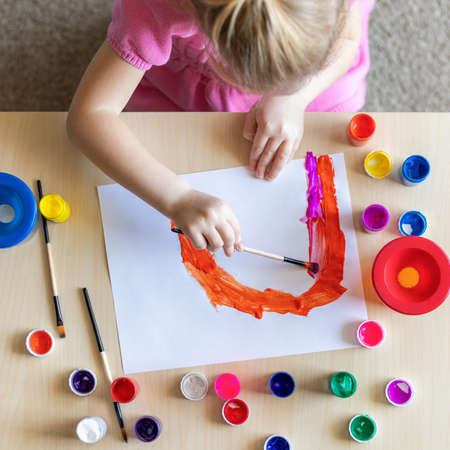 Little girl painting rainbow on paper with colorful paints sitting at the table at homeの写真素材