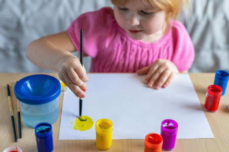 Little smiling girl painting on paper with colorful paints sitting at the table at home