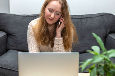 Young woman working, sitting on the couch with laptop, talking on the phone. Business, working from home conceptの写真素材