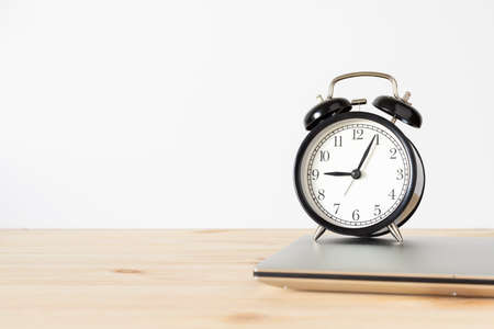 Classic vintage black alarm clock and laptop on wooden table with white wall background and copy space for text. Time and work concept.の写真素材