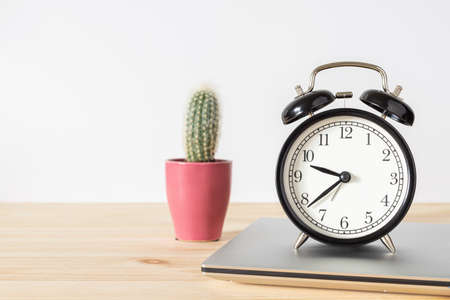 Classic vintage black alarm clock, laptop and small potted cactus on wooden table with white wall background and copy space for text. Time and work concept.の写真素材