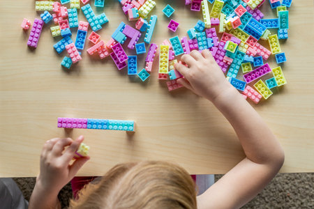 Little beautiful girl playing with toy plastic building blocks, sitting at the table. Small child...
