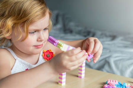 Little beautiful girl playing with toy plastic building blocks, sitting at the table. Small child...