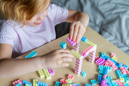 Little beautiful child playing with toy plastic building blocks, sitting at the table. Small girl...