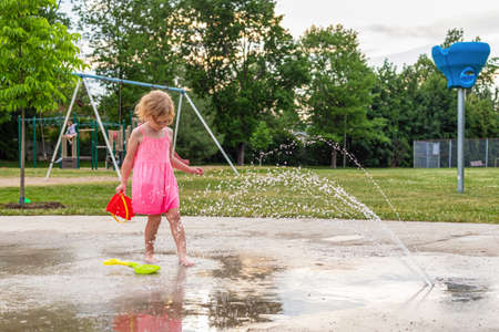 Little child playing with water and toys at splash pad in the local public park during hot summer day. Small beautiful girl in pink dress having fun at fountain playground.の写真素材