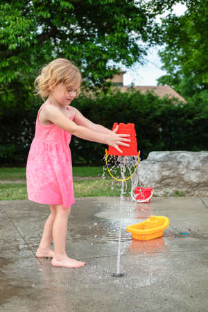 Little child playing with water and toys at splash pad in the local public park during hot summer day. Small beautiful girl in pink dress having fun at fountain playground.の写真素材