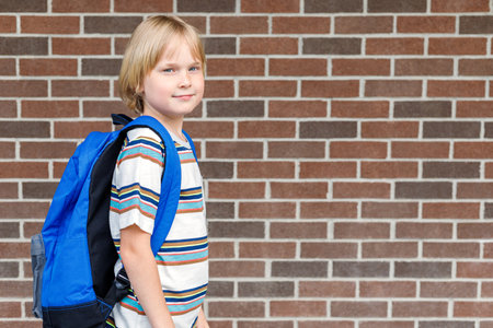 Beautiful child near school building at the schoolyard against brick wall with copy space. Little blond student carrying blue backpack. Back to school concept.の写真素材
