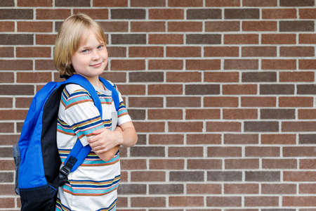 Beautiful smiling child near school building at the schoolyard against brick wall with copy space. Little blond student carrying blue backpack. Back to school concept.の写真素材