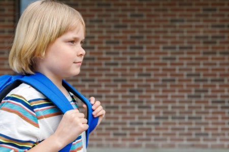Beautiful child near school building at the schoolyard against brick wall. Small blond student carrying blue backpack. Back to school concept with copy space for text.の写真素材