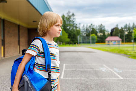Beautiful happy child near school building at the schoolyard. Little smiling student carrying blue...