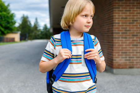 Beautiful small child near school building at the schoolyard. Little blond student with blue backpack.の写真素材