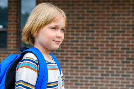 Beautiful child near school building at the schoolyard against brick wall. Small blond student carrying blue backpack. Back to school concept with copy space for text.の写真素材