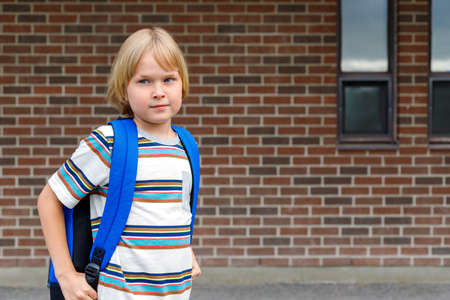 Cute kid near school building at the schoolyard against brick wall with copy space. Little blond schoolboy carrying blue backpack. Back to school concept.の写真素材