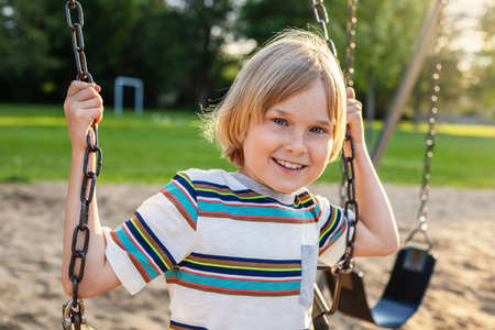 Happy smiling child sitting on swing at public park playground near school on a sunny summer day. Littke kid swinging, having funの写真素材