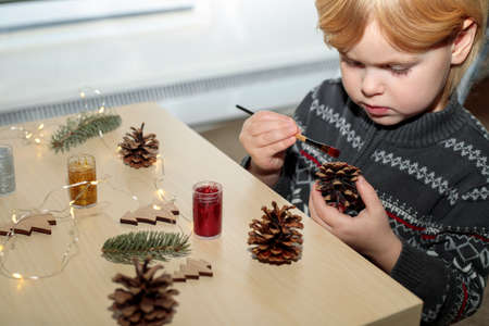 Christmas kids craft activities at home. Little child painting pine cones and wooden Christmas tree decorations at home. Holiday DIY concept.の写真素材