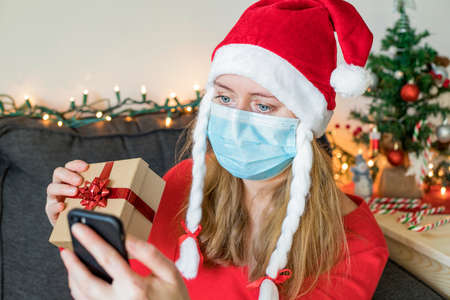 Christmas online congratulations. Woman in mask making video call over smart phone on Christmas Eve, greeting with holidays, holding gift box. Christmas celebration on distance concept.の写真素材