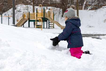 Small child making snowman, playing with snow near playground on snowy cold winter day. Girl wearing snowsuit outdoors. Fun outdoor activity for kids and family.の写真素材