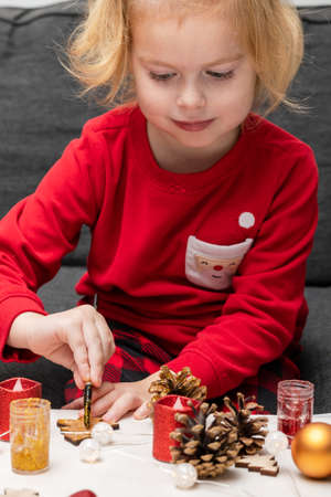 Small girl doing crafts, wearing red santa pajamas, sitting at table. Little child painting pine cones and wooden Christmas tree decorations at home. Holiday DIY concept for children.の写真素材