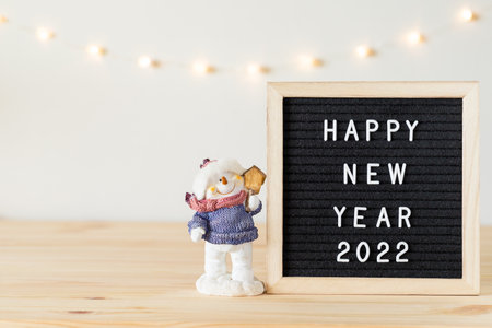 Happy new year 2022 greetings on black felt letter board with toy snowman on wooden table and holiday lights on wall. Copy space for textの写真素材