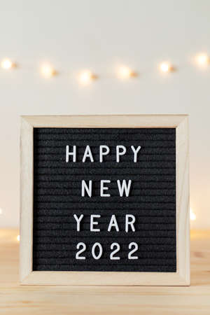 Happy new year 2022 greetings on black felt letter board on table with holiday decorative lights on white wall.の写真素材